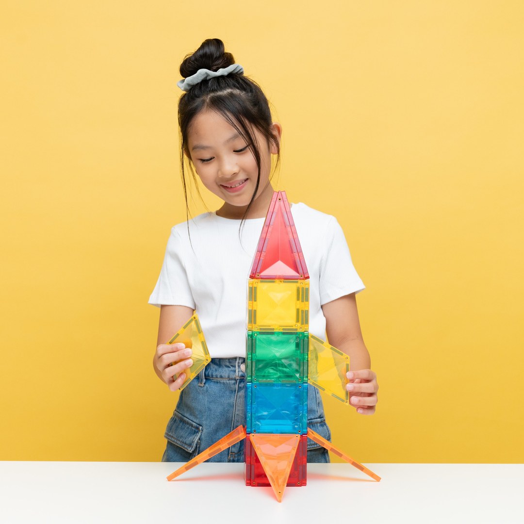Smiling girl building a rainbow rocket from CONNETIX magnetic tiles on a yellow background