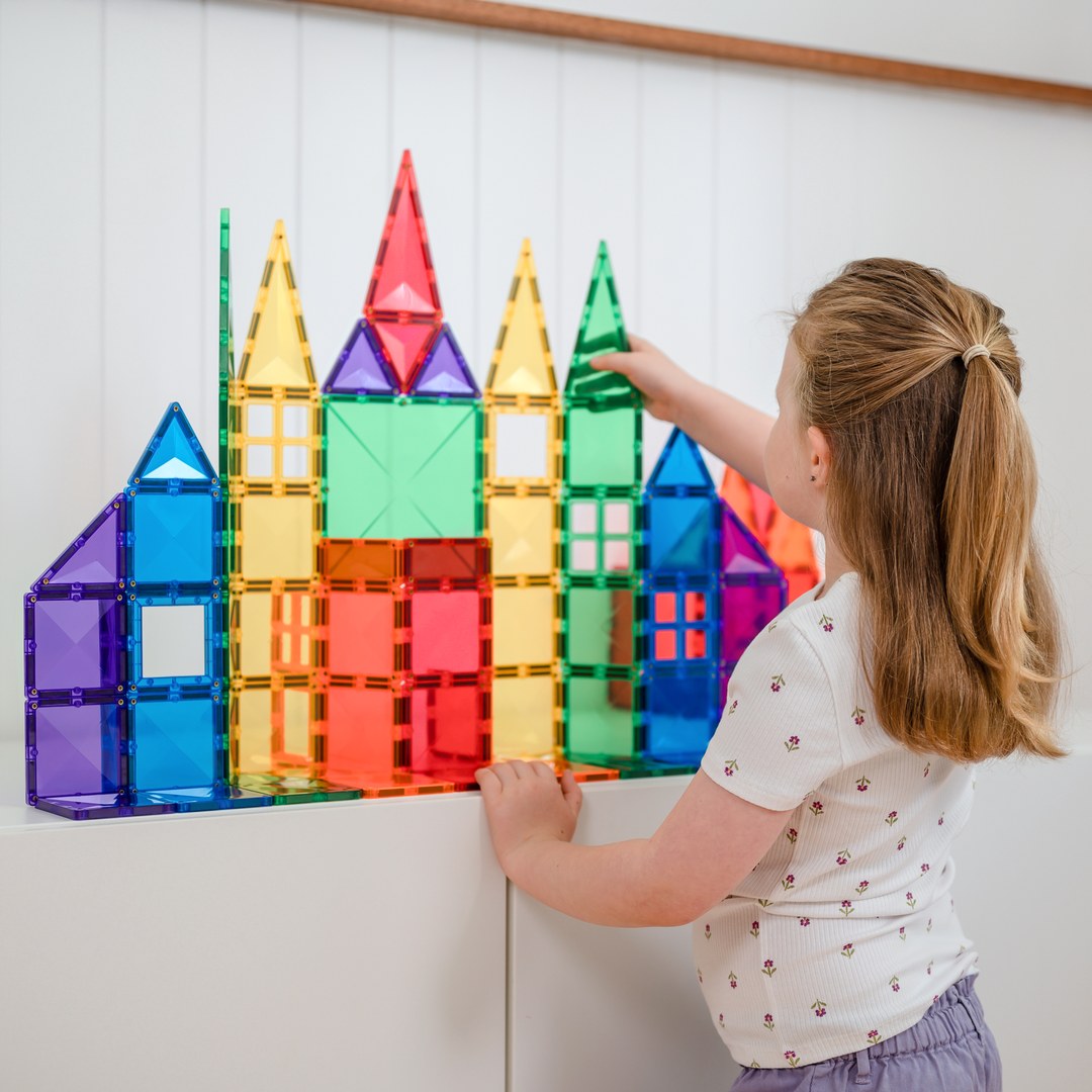 Girl adding a green triangle to a rainbow castle built from CONNETIX magnetic tiles