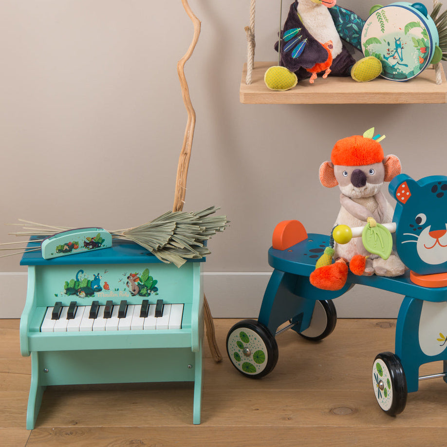 Collection of children's toys including a toy piano, tricycle, and guitar on a wooden floor.