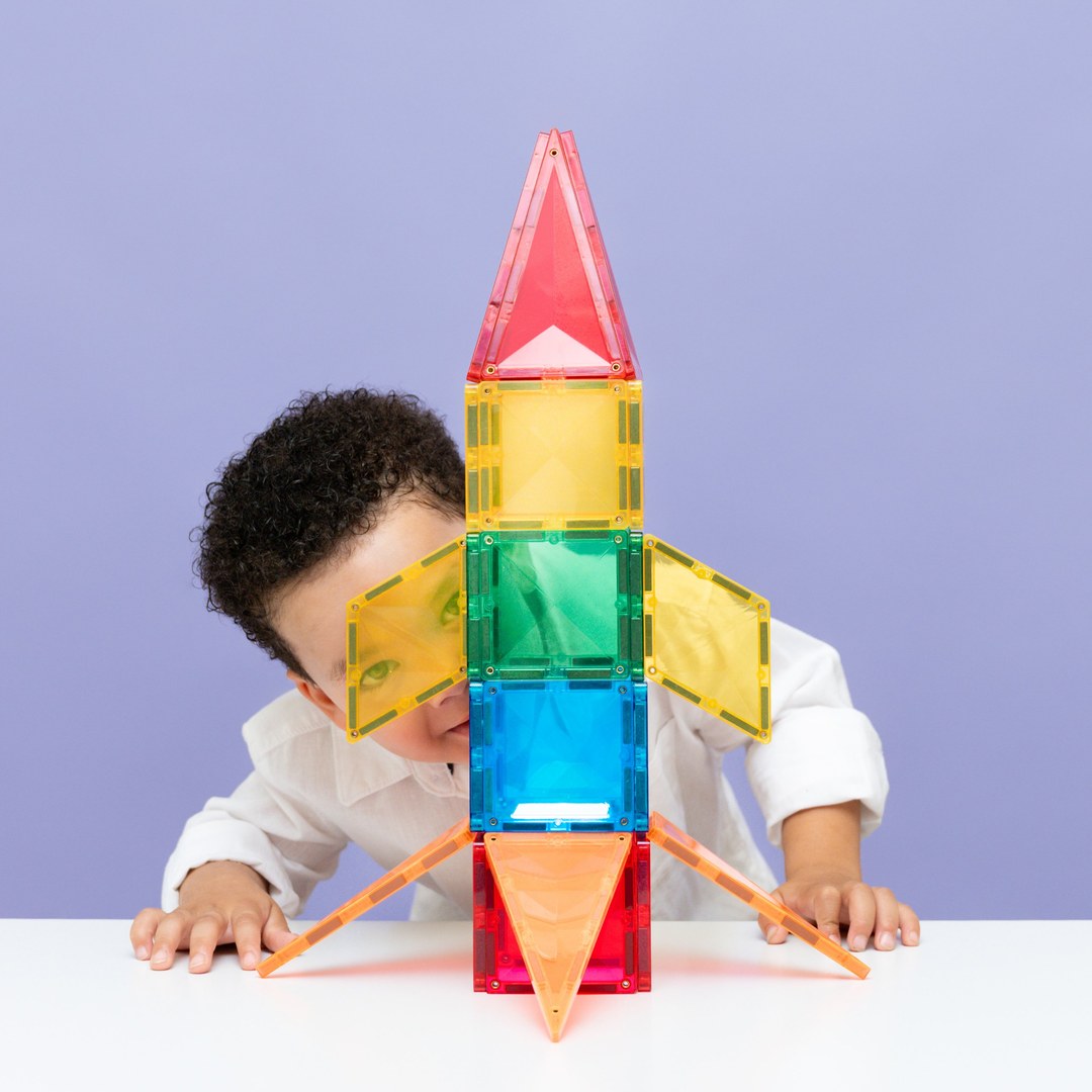 Young boy peeking through a CONNETIX magnetic tile rocket with yellow wings and orange fins
