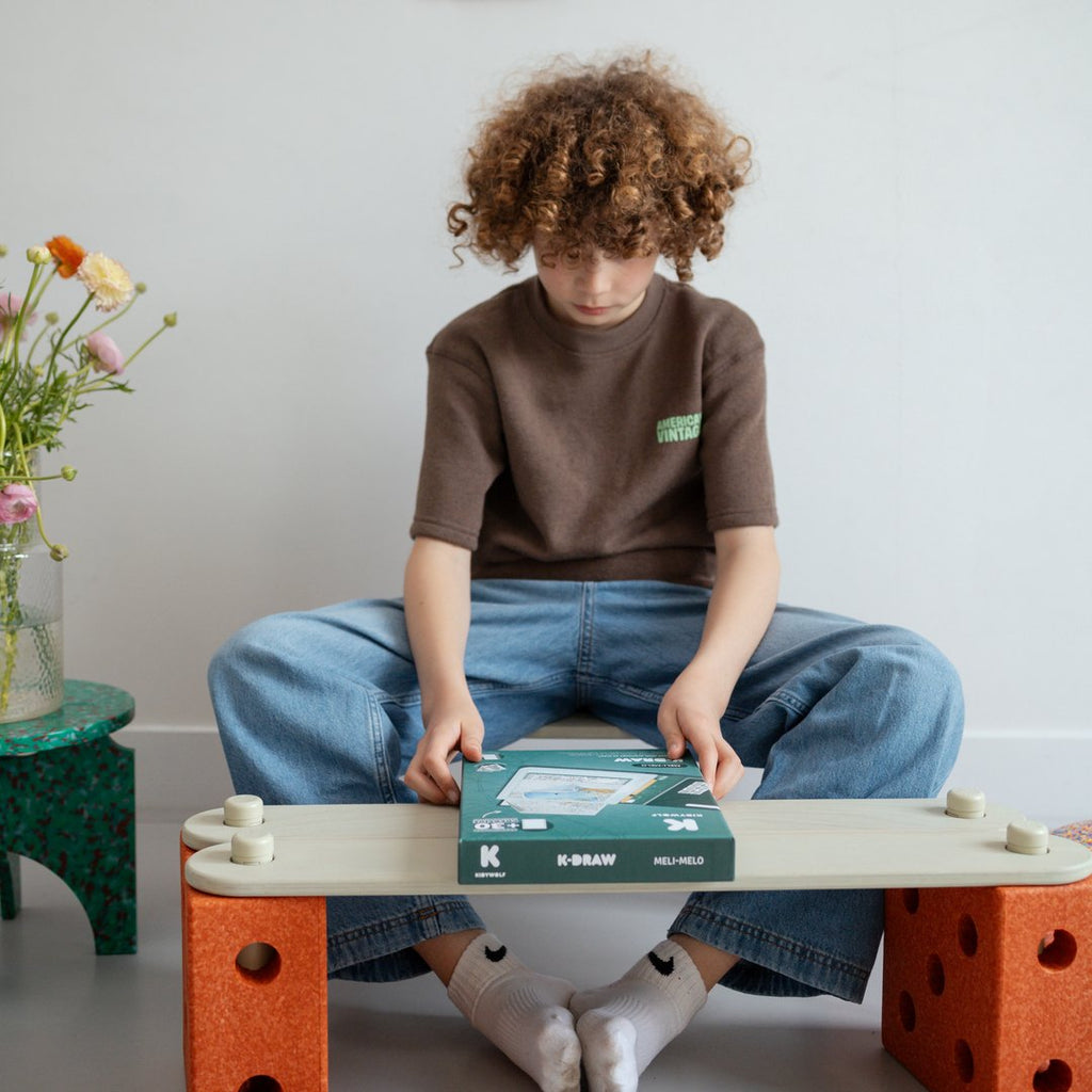 Child sitting cross-legged on a bench opening the K-DRAW Meli Melo box