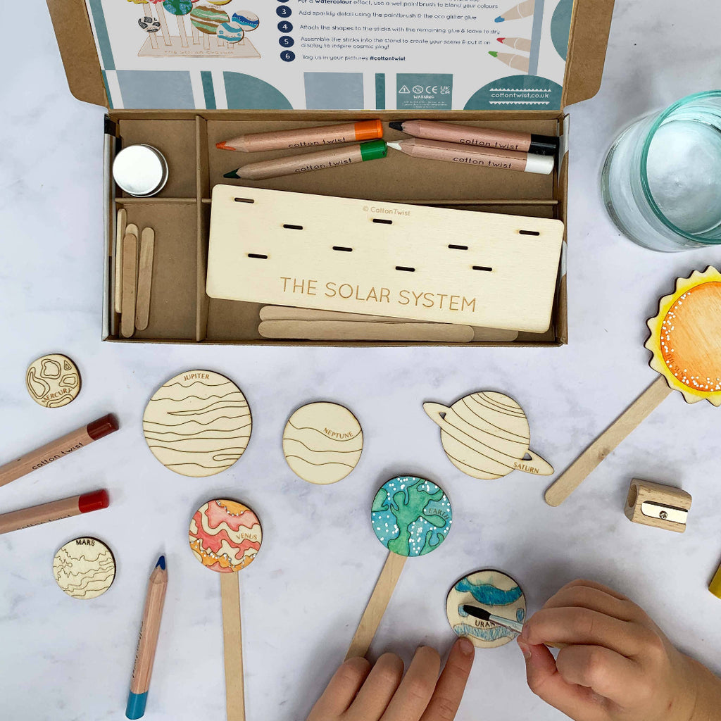Solar system model kit with wooden planets, solar system-themed pencils, and a box labeled 'The Solar System' on a light gray background.