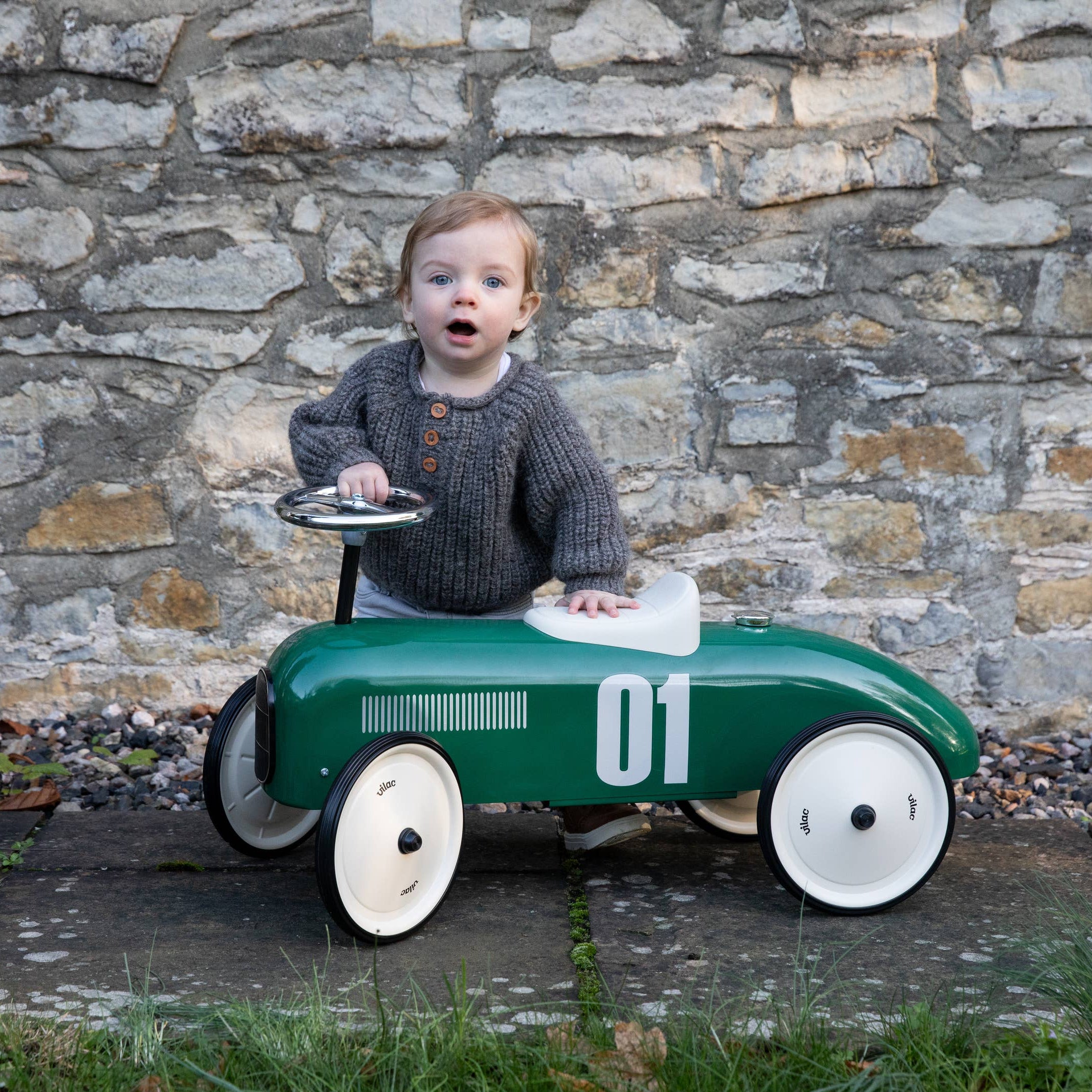 Child sitting in a green toy car with white wheels against a stone wall.