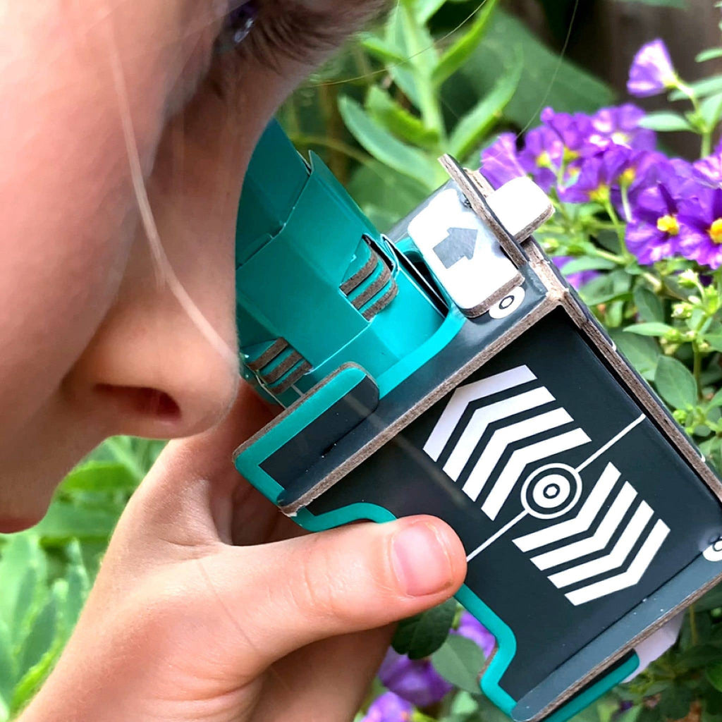Person holding microscope lense, in garden surrounded by green leaves and purple flowers. examining flower 