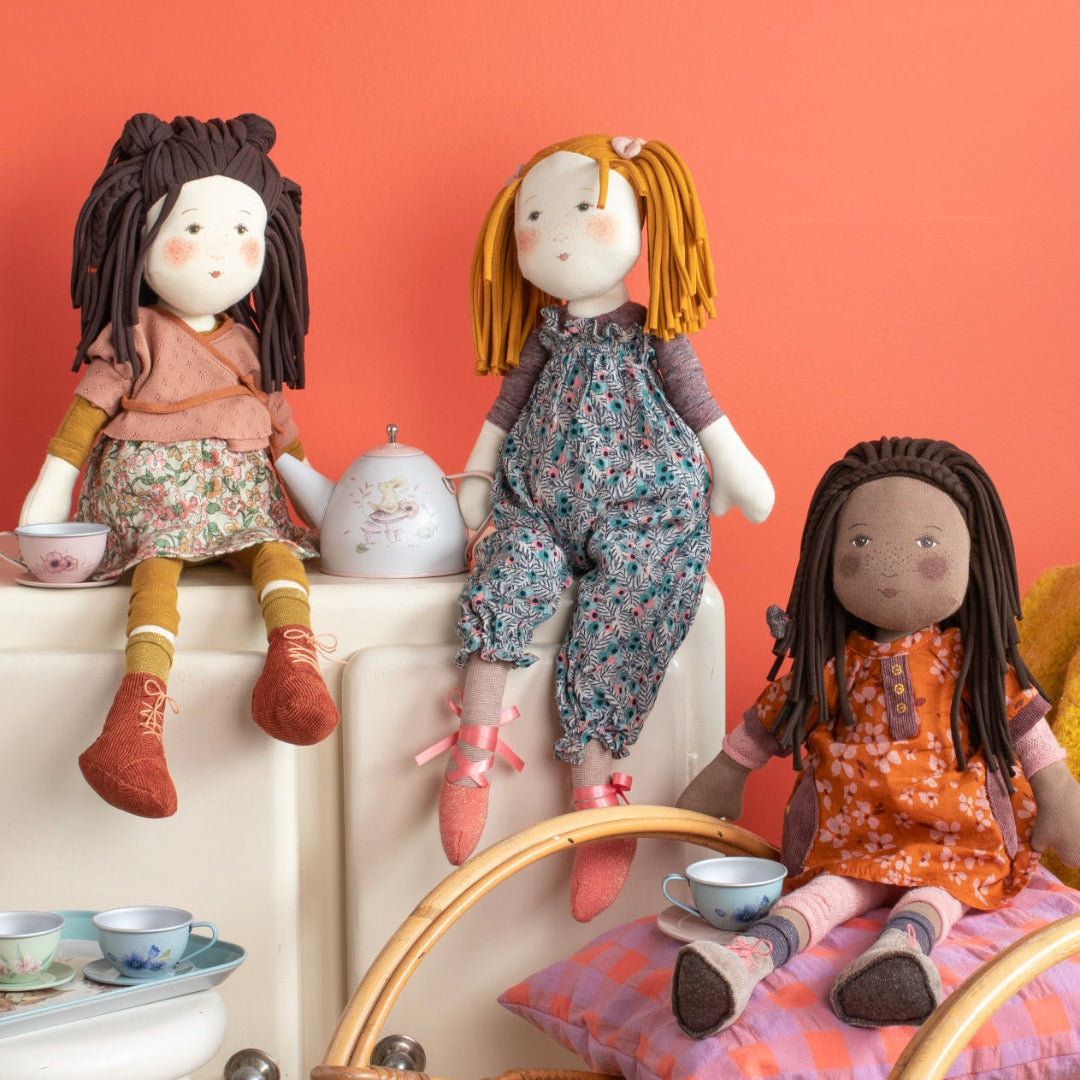 Three dolls with different hairstyles and outfits sitting on a chair and a small table against an orange wall.