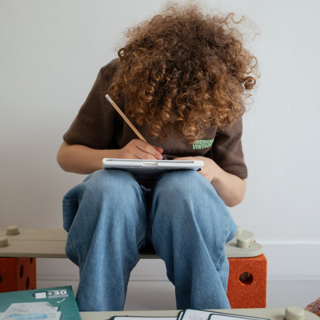 Child sitting on a bench tracing a drawing on the KIDYDRAW Pad resting on their knees