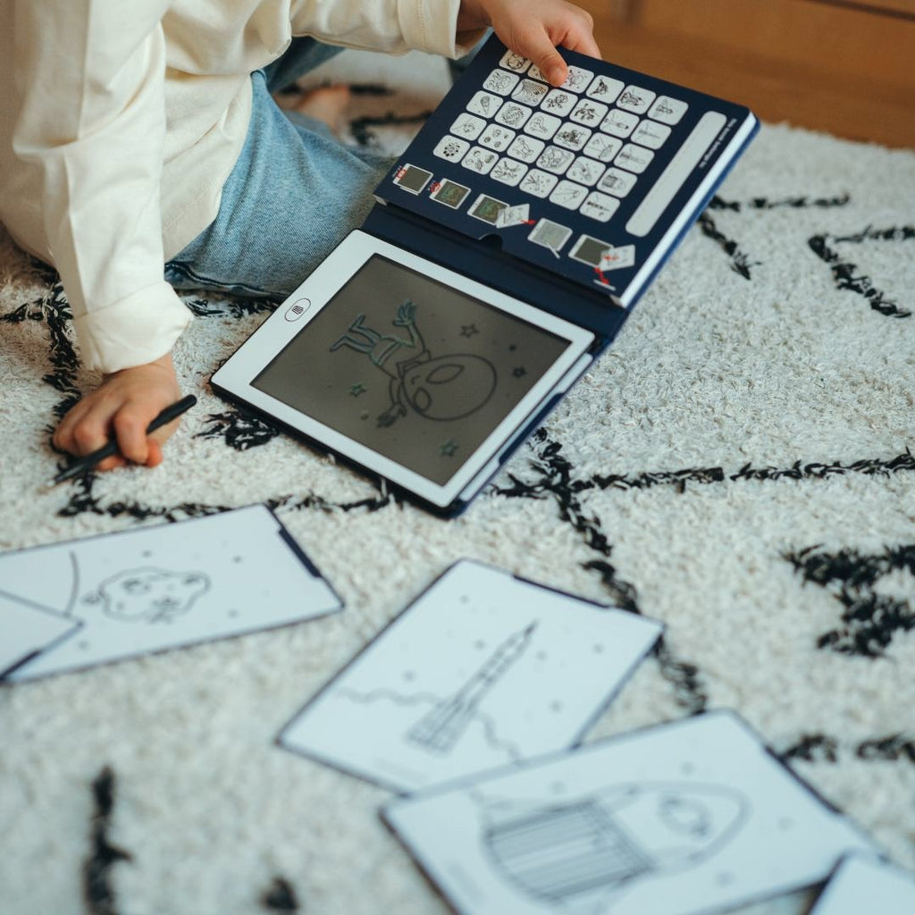 Child kneeling on a rug with the navy KIDYDRAW MINI showing an alien drawing, surrounded by Galaxy rocket and asteroid cards