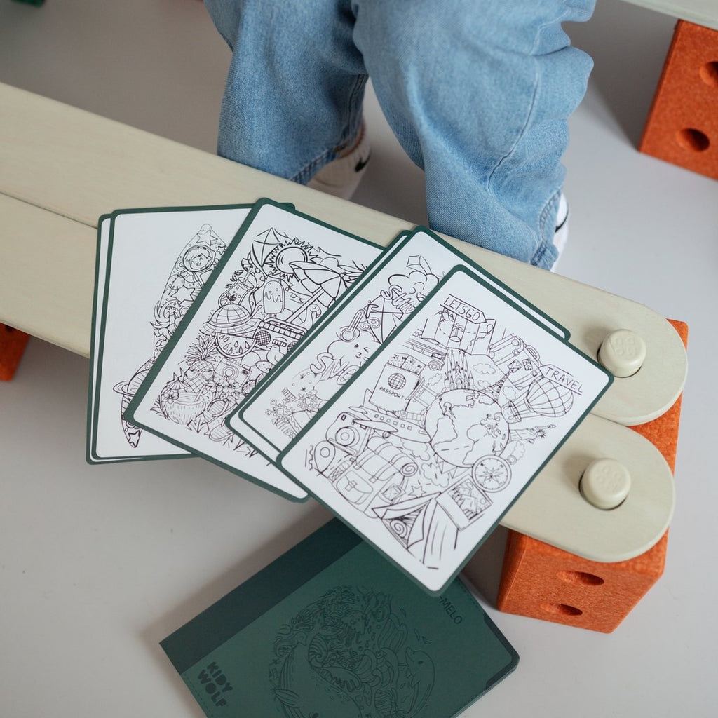 Meli Melo illustration sheets and the green KIDYDRAW booklet laid on a bench while a child traces