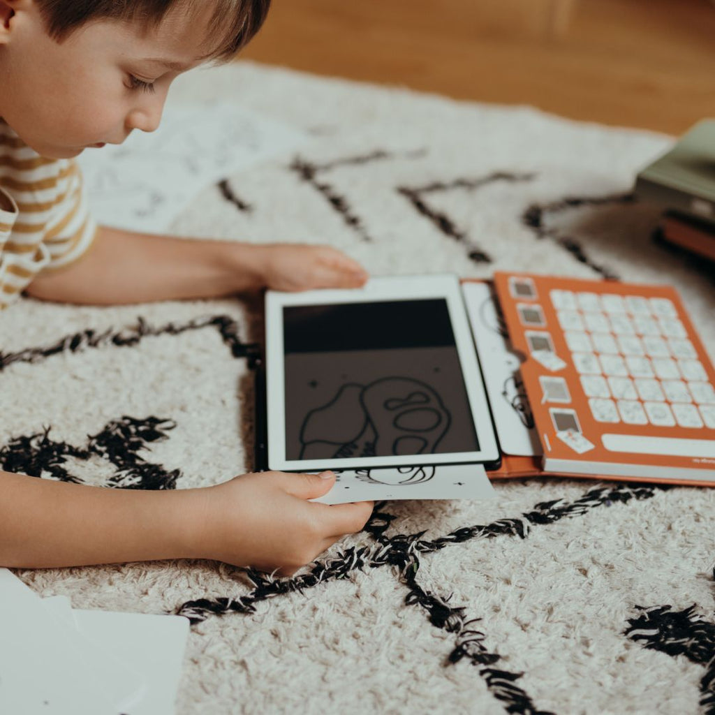 Young boy lying on a rug sliding a dinosaur card into the KIDYDRAW MINI tablet beside the orange Dinosaurs card holder