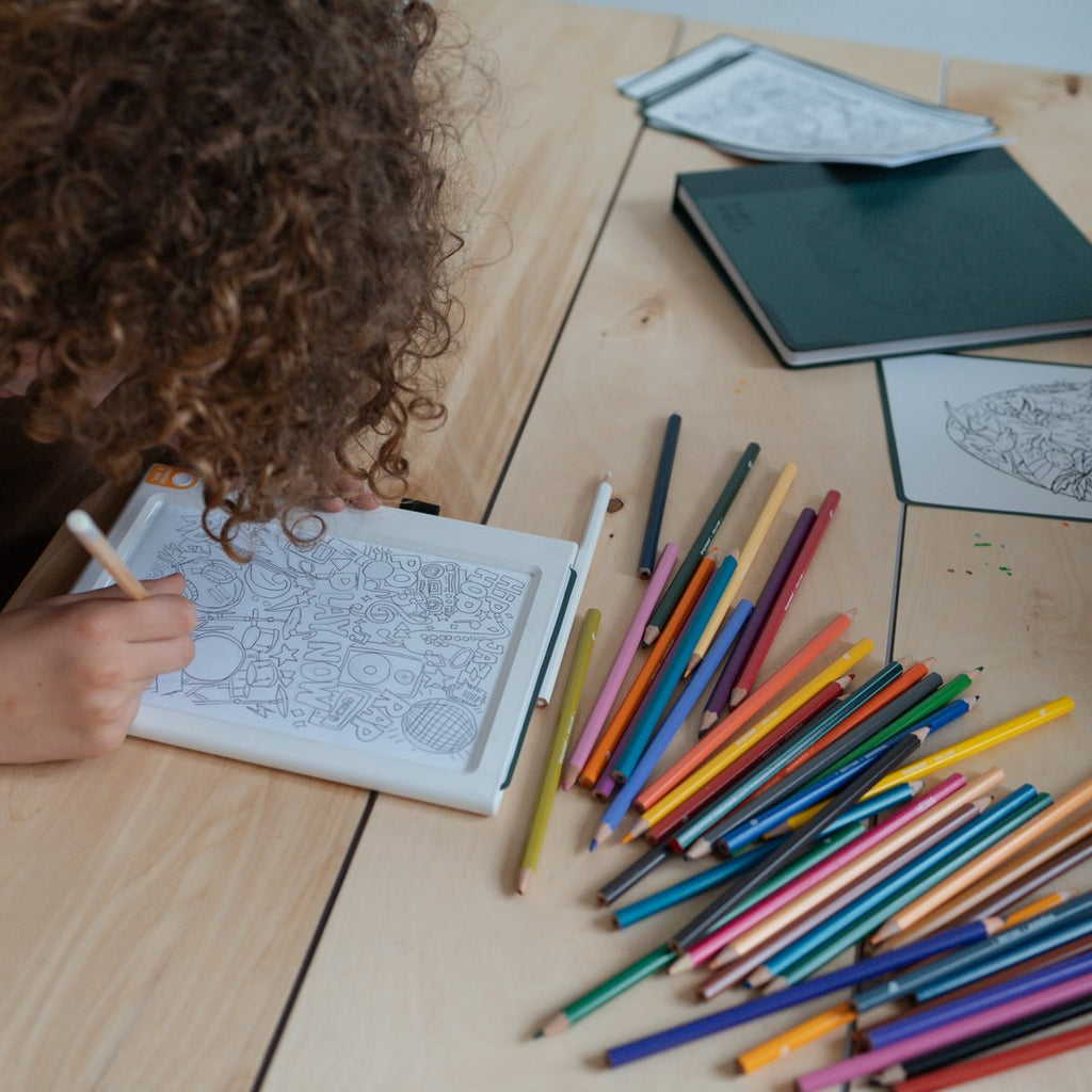 Child tracing at a wooden table with the KIDYDRAW Pad and a pile of coloured pencils