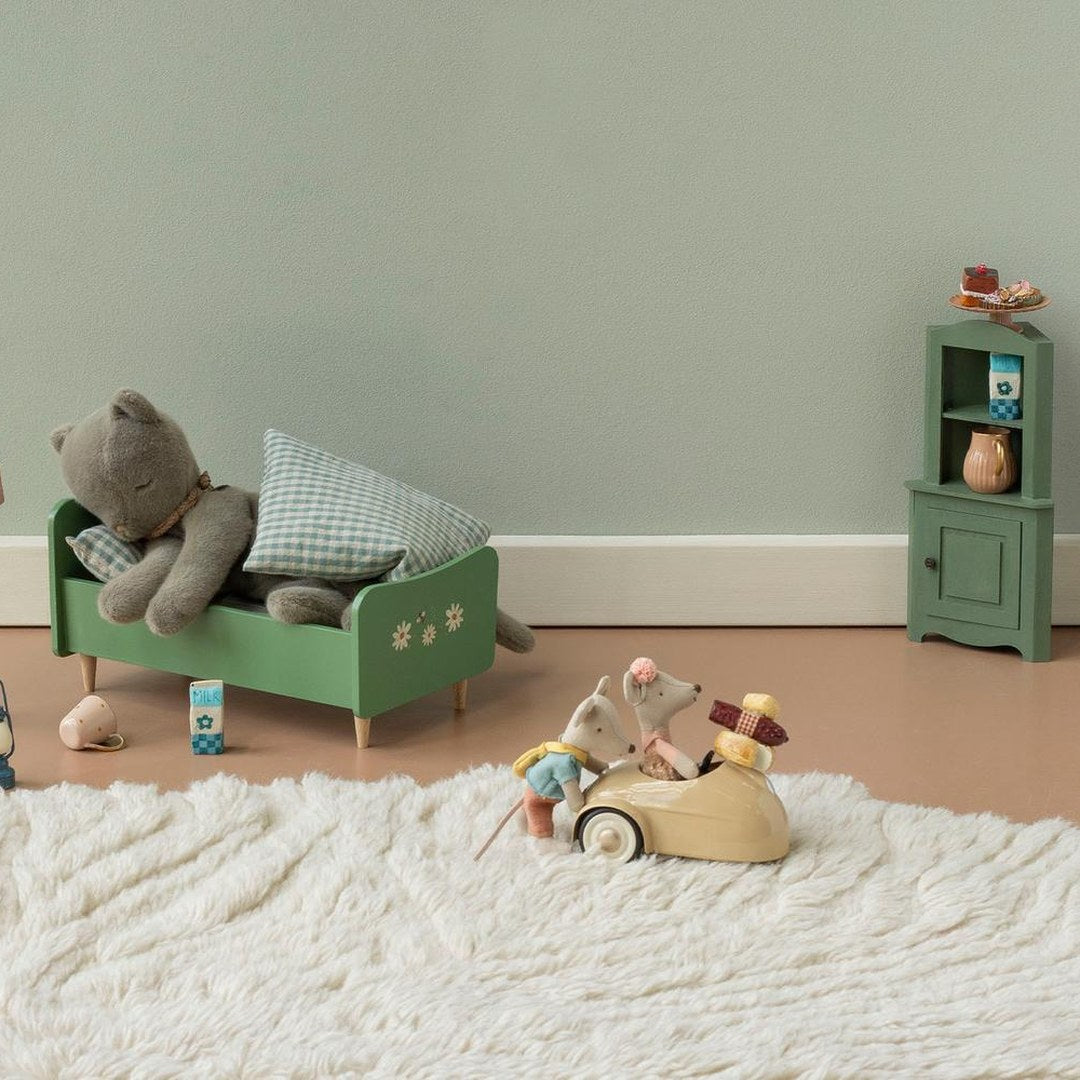 Maileg mice driving a cream mouse car across a fluffy rug past a sleeping cat in a green bed