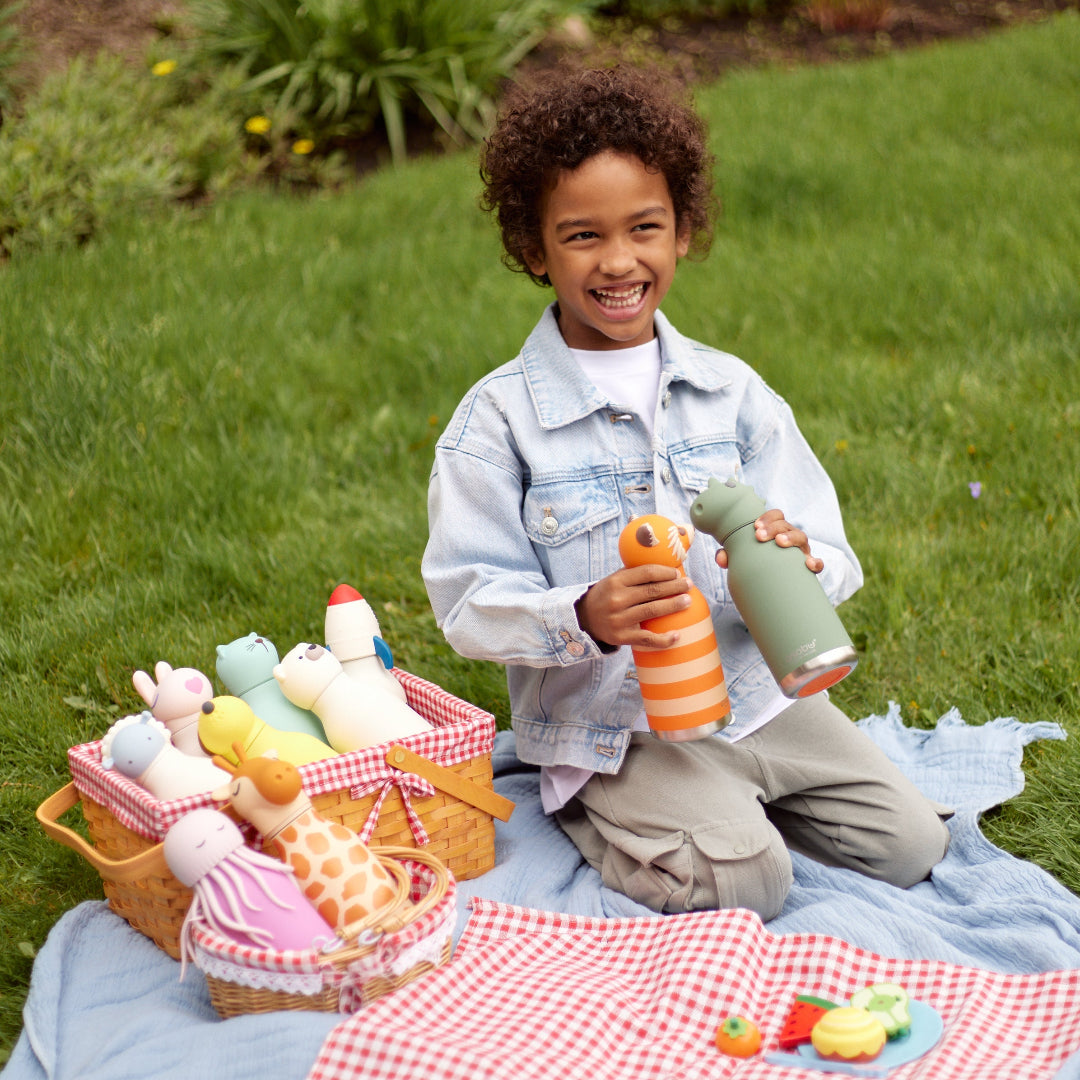 A smiling young child kneels on a blue picnic blanket in a grassy backyard, holding a green bottle and an orange-and-white striped bottle. A woven basket filled with plush toys sits beside the blanket, with a red-and-white check picnic cloth and toy food in front.