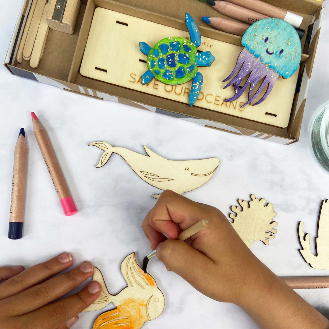 Children's hands coloring wooden marine animal cutouts with markers, including a turtle, jellyfish, whale, and coral.
