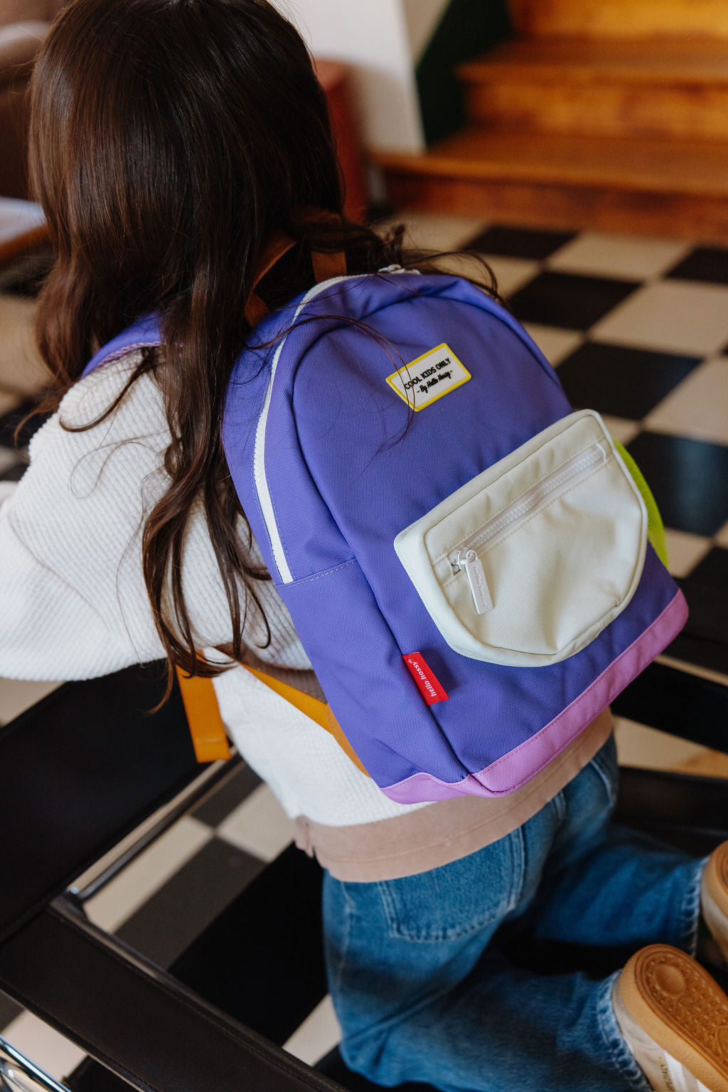 A child wearing a purple backpack with a white front pocket, photographed from behind in an indoor setting with a black-and-white checkered floor.
