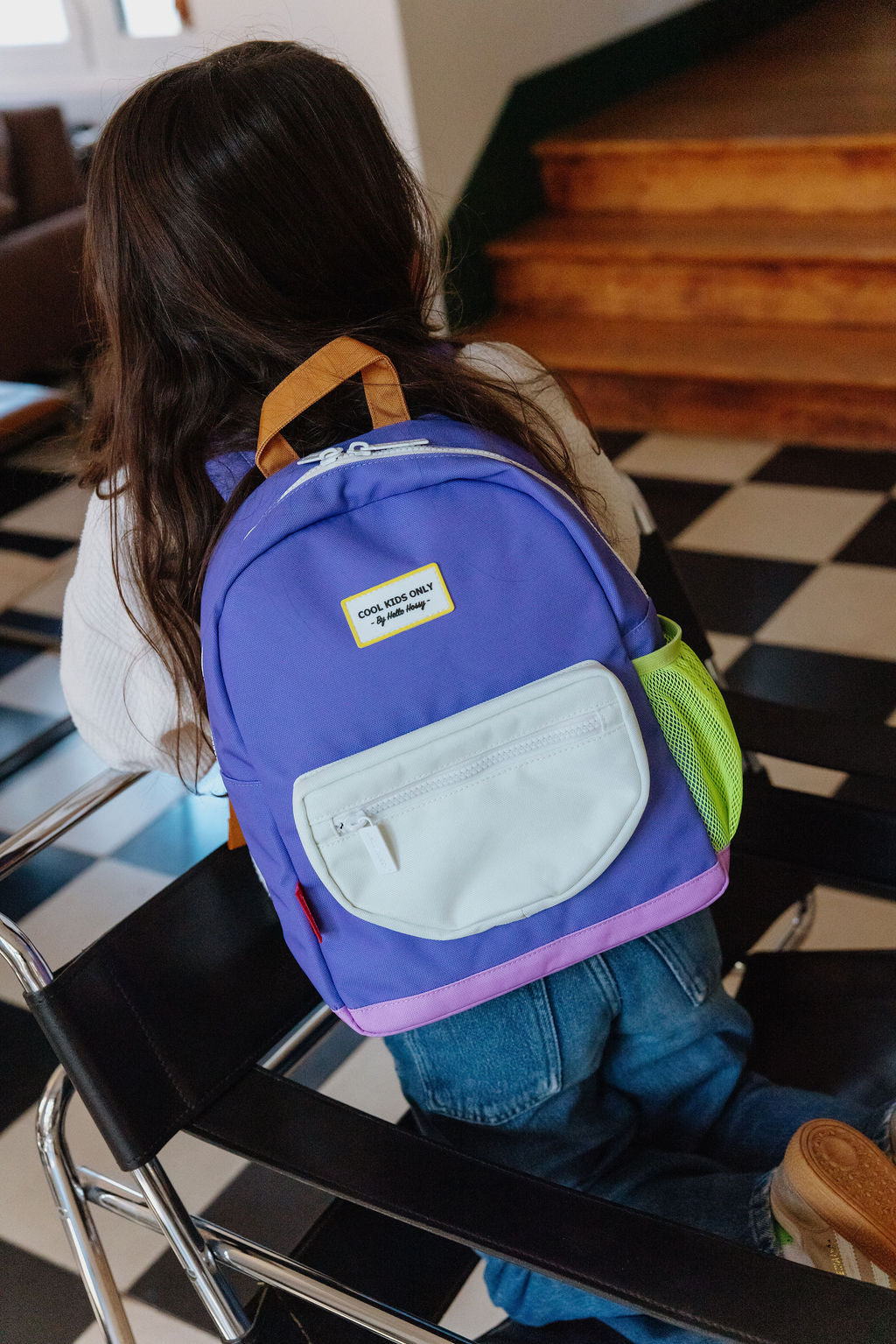 A child seen from behind wearing a purple backpack with a white front pocket and tan shoulder straps, sitting in a chair in an indoor setting with a black-and-white checkered floor and wooden stairs in the background.