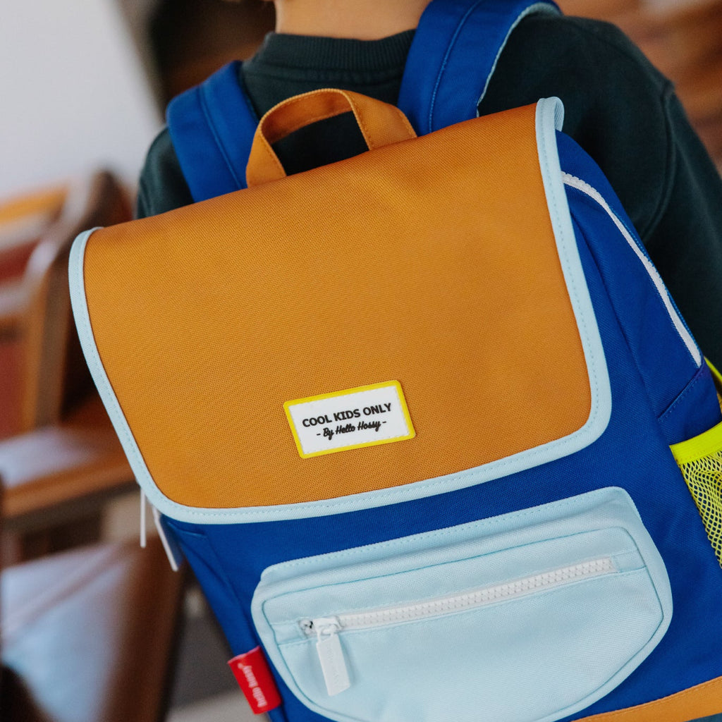A child viewed from behind wearing a blue, mustard yellow, and light blue school backpack with a 'Cool Kids Only' brand patch on the front.