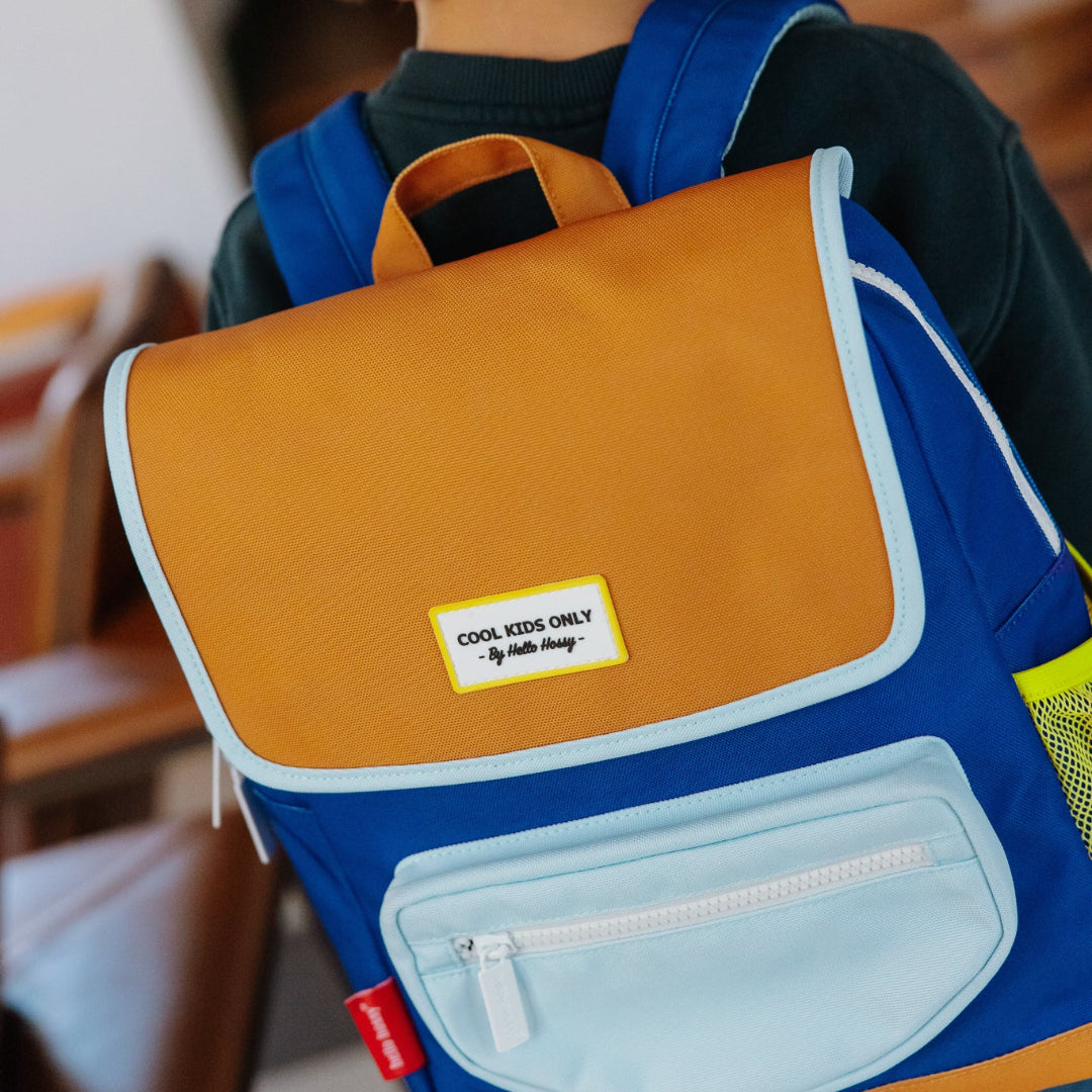A child viewed from behind wearing a blue, mustard yellow, and light blue school backpack with a 'Cool Kids Only' brand patch on the front.