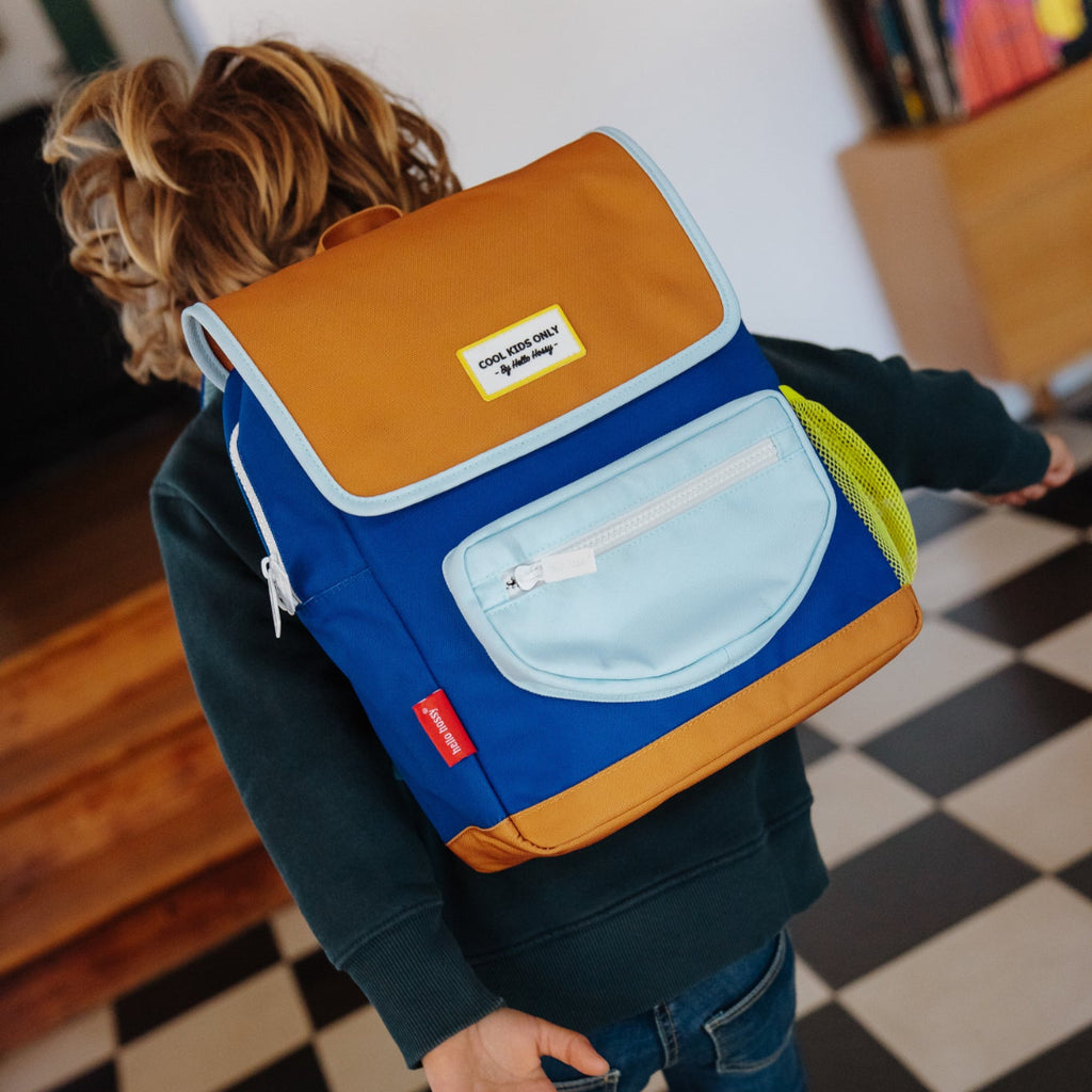 A child wearing a dark green sweatshirt, blue jeans, and sneakers, shown from behind while wearing a colorful backpack. The backpack features a mustard yellow top flap, a light blue front pocket, a medium blue body, and a yellow-green side pocket.