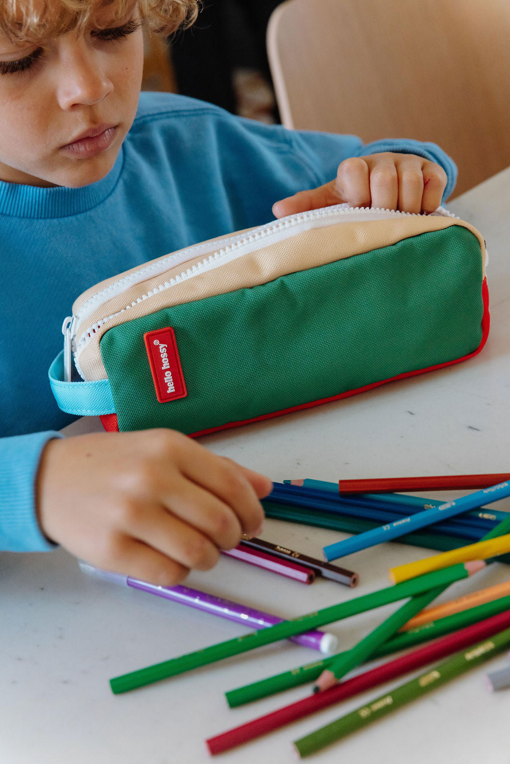 A child in a blue sweatshirt looks down at a green, red, and beige zippered pencil case while sitting at a white table with colored pencils scattered in front.