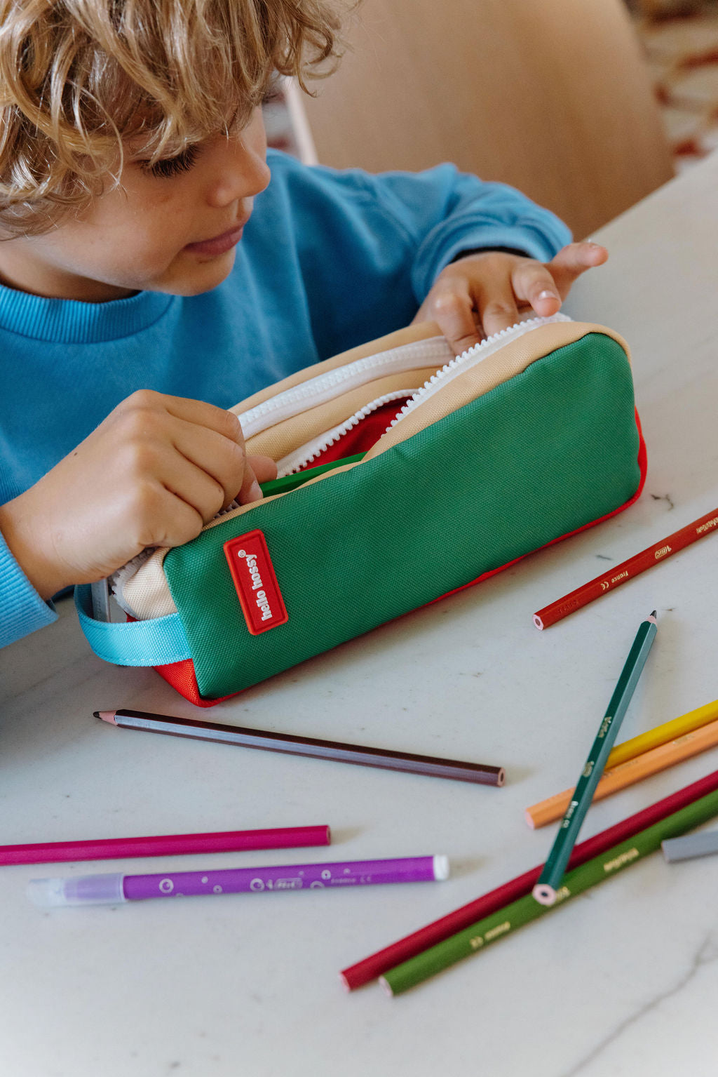 A child in a blue sweater opens a green and red pencil case on a white table, with colored pencils scattered around.