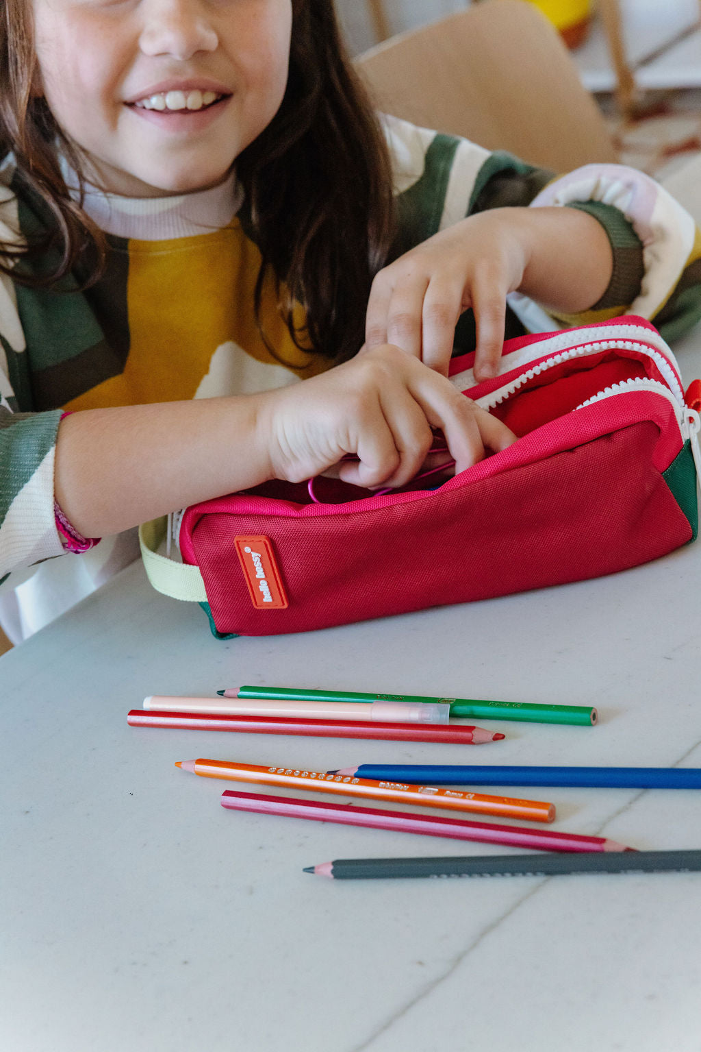 A child's hands and smiling face are visible as they open a red fabric pencil pouch on a light gray table. Several colored pencils are scattered in front of the pouch.