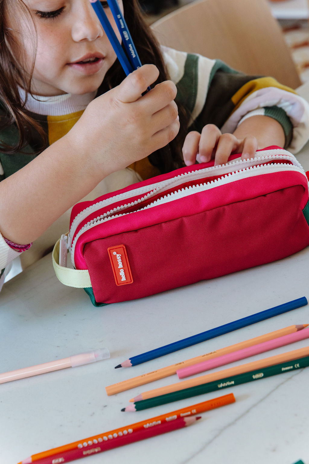 A child holding blue pencils over a bright red zippered pencil case on a white table, with coloured pencils scattered nearby. 