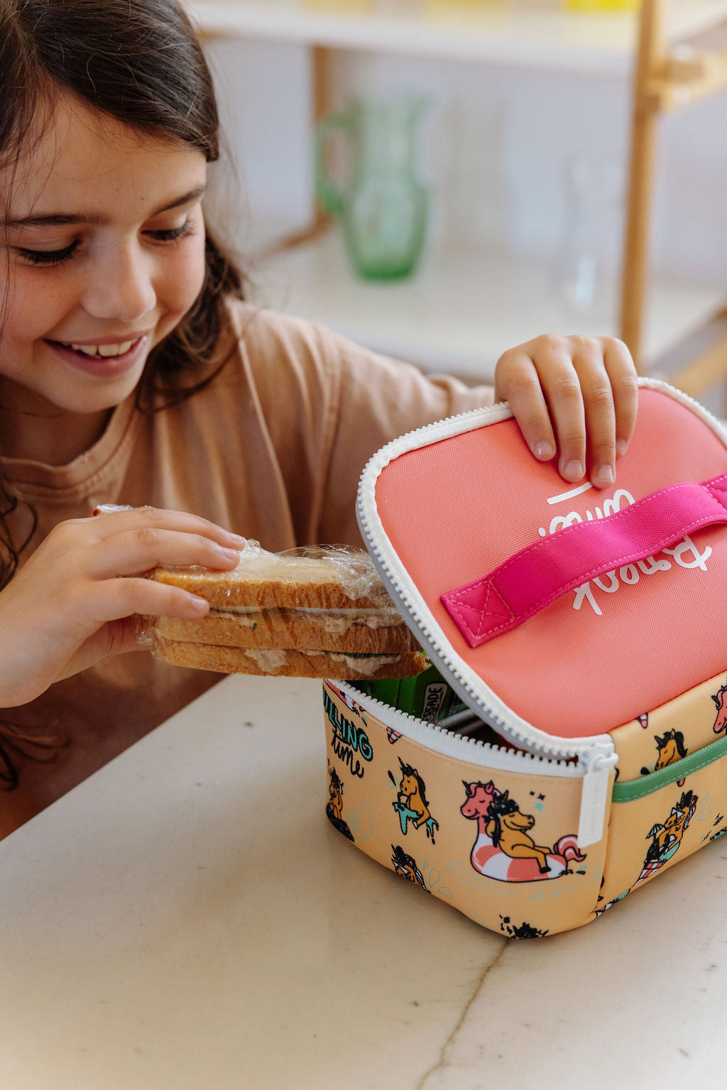 Child holding a pink lunch box with a cartoon design on a table.