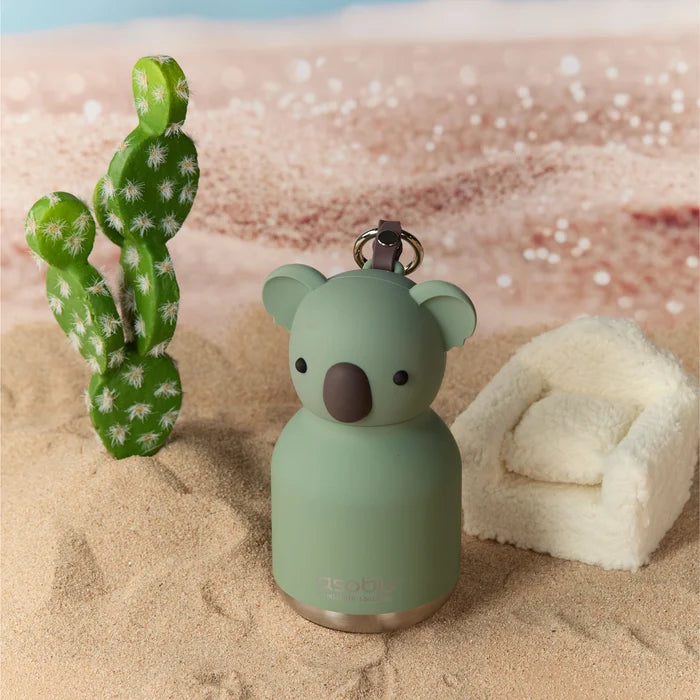 Green koala-shaped bottle keychain on a sandy surface with a cactus and towel in the background
