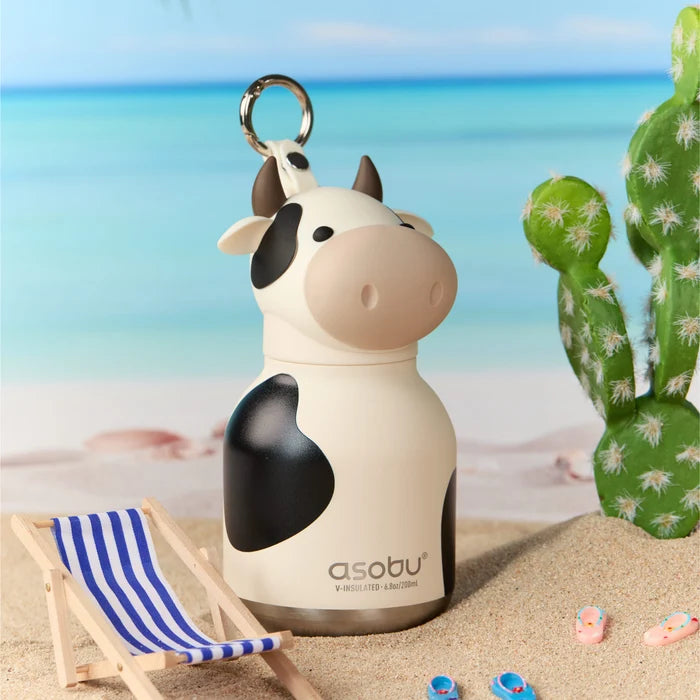 Cow-shaped water bottle with 'asobu' branding on a beach setting with a cactus and striped chair.