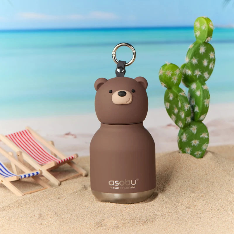Brown bear-shaped water bottle with a keychain on a sandy beach with a cactus and deck chair in the background.