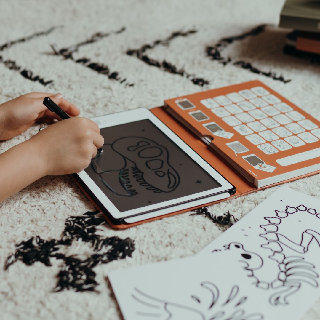 Child tracing a T-rex skull onto the KIDYDRAW MINI tablet with the stylus, orange card holder open beside a dinosaur skeleton card