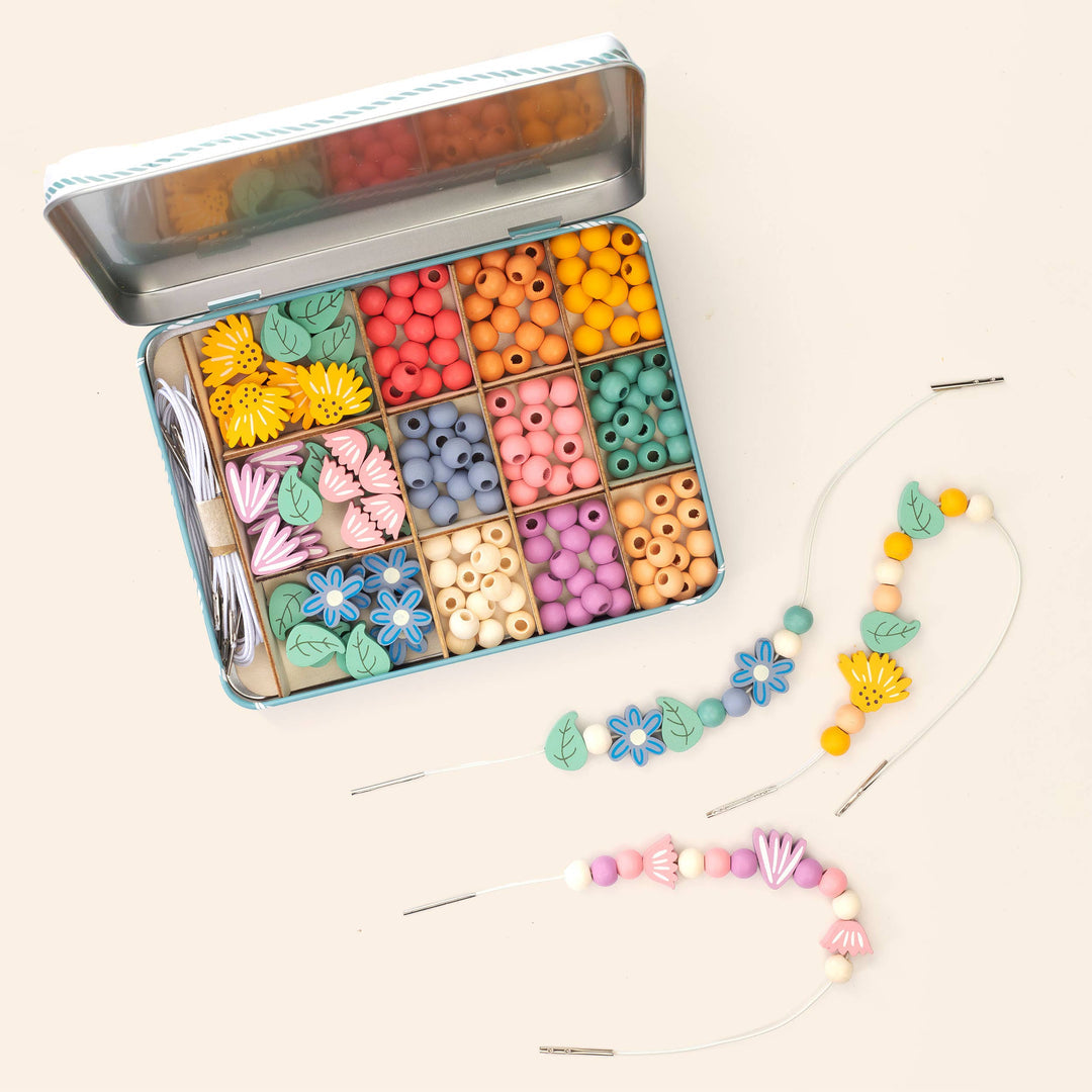 Open tin of colourful wooden flower, leaf and round beads with two part-threaded bracelets on a cream background
