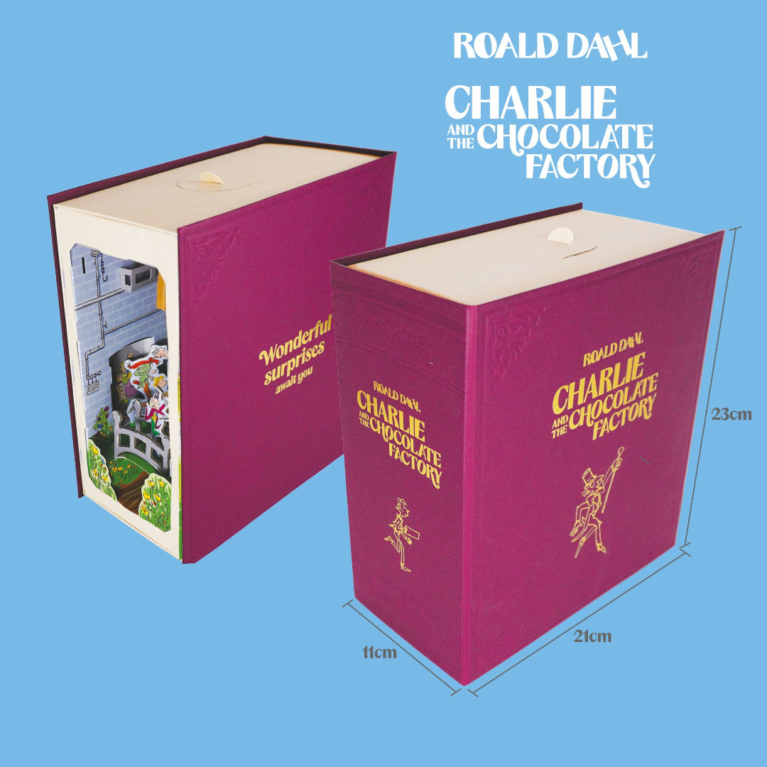 A Roald Dahl 'Charlie and the Chocolate Factory' surprise box shown in two views on a light blue background, with printed dimensions labeled 23cm height, 11cm width, and 21cm depth. One box is closed in magenta with gold lettering, and the other is open to reveal a colorful chocolate factory scene inside.