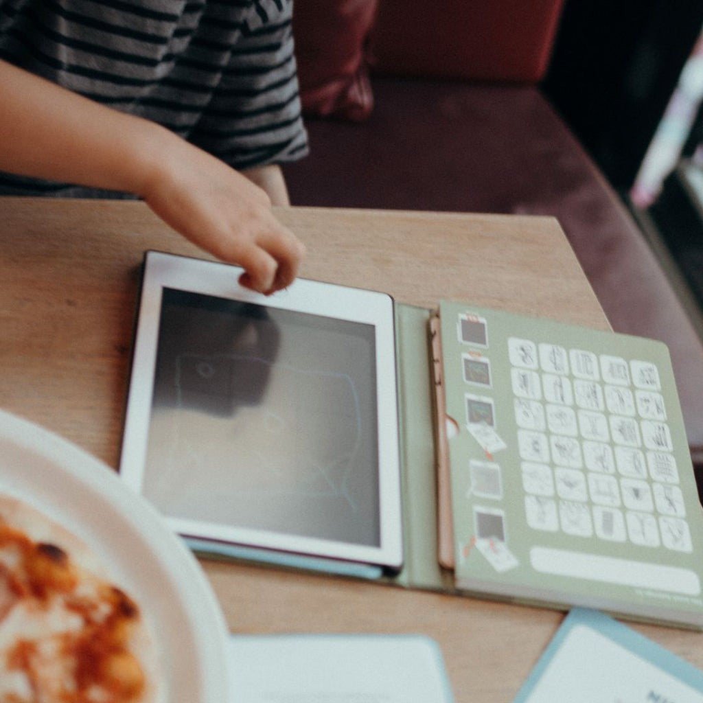 Child's hand reaching towards a tablet on a wooden table with a plate of food.