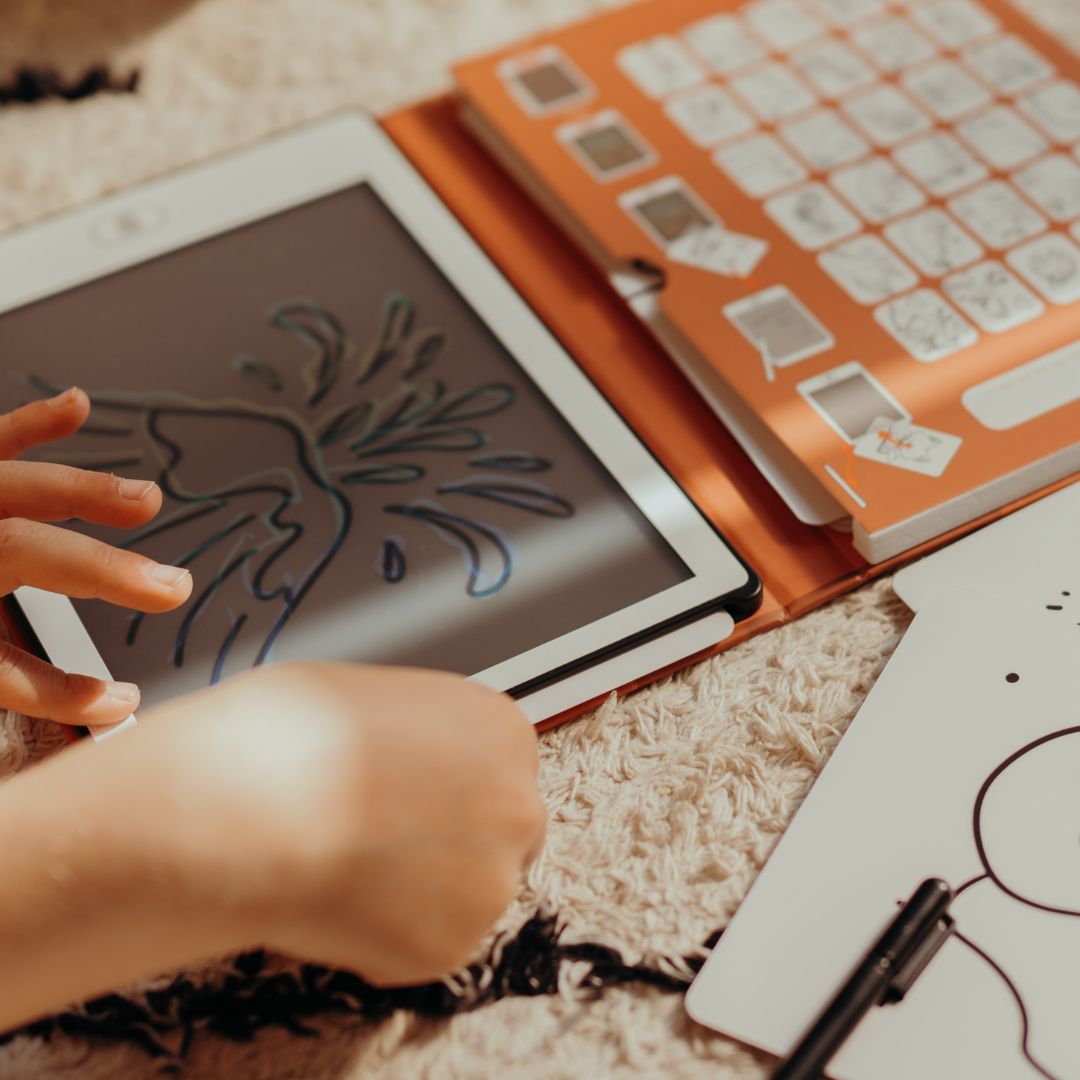 Close-up of a child's hand drawing a volcano outline on the KIDYDRAW MINI screen next to the orange Dinosaurs card holder
