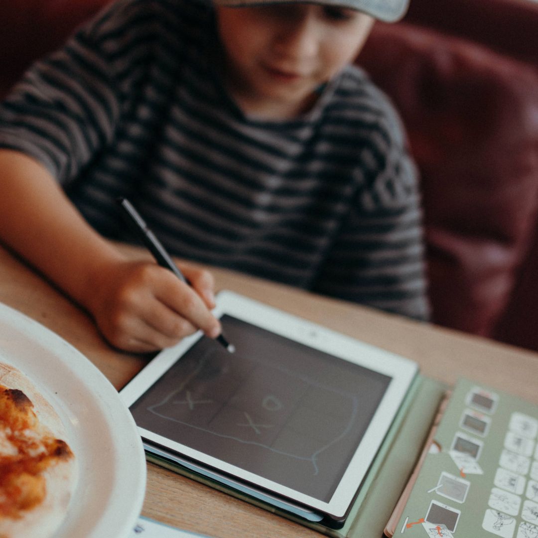 Child using a stylus on a tablet with a plate of food and book on a table