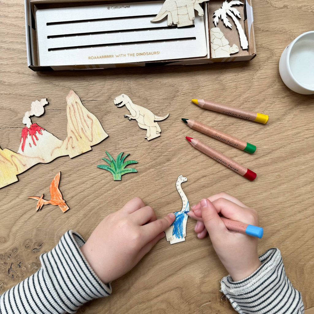Children's hands coloring dinosaur figures on a wooden table with crayons and a box of figures.