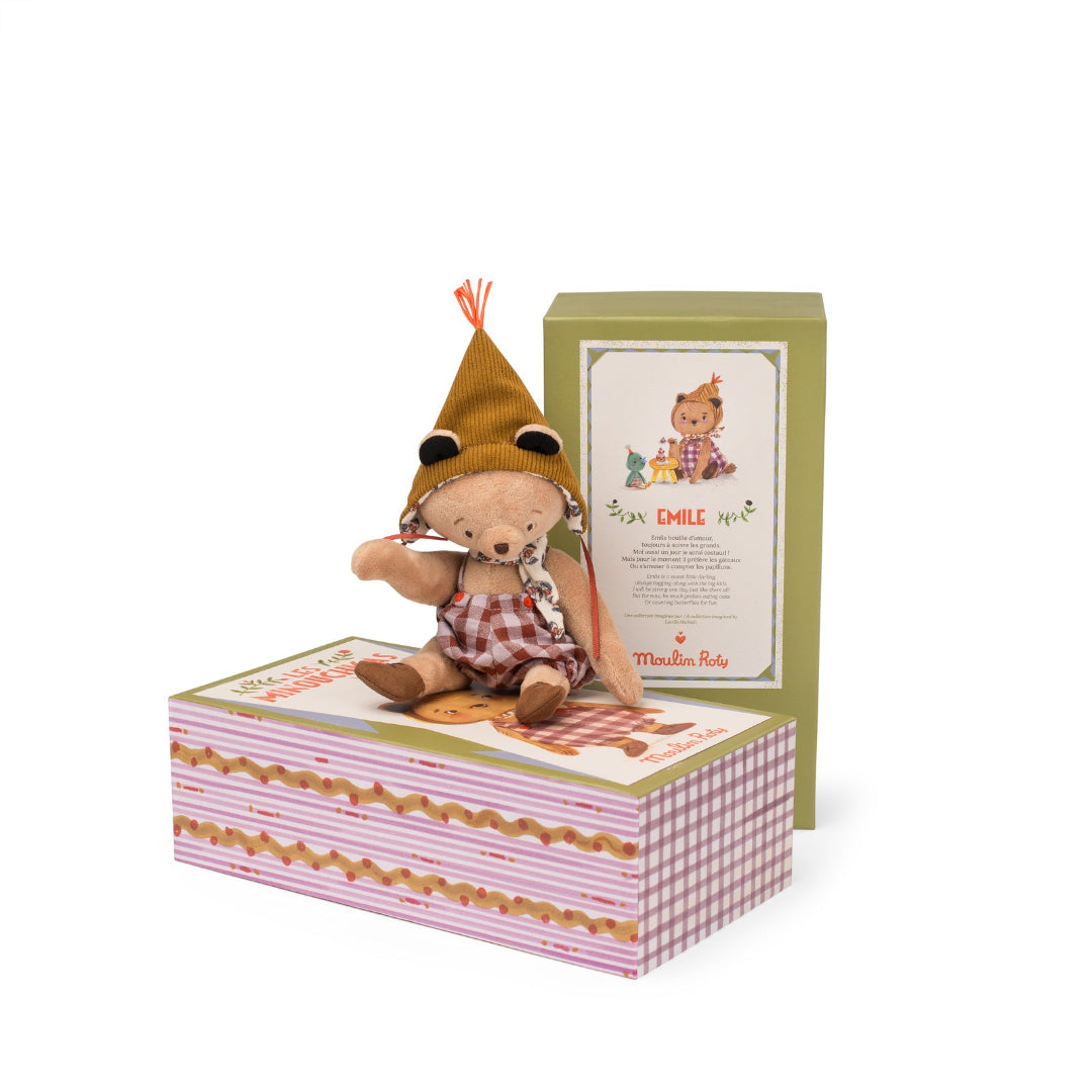 Small teddy bear in a checkered outfit with a hat, sitting on a decorative box with a card, on a white background.
