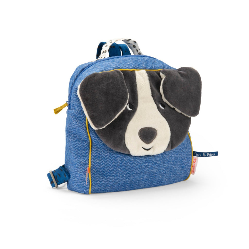 A blue denim-textured dog face backpack by Skip Hop, featuring a plush black and white dog face on the front with floppy ears, yellow piping details, and a small brand tag on the side.