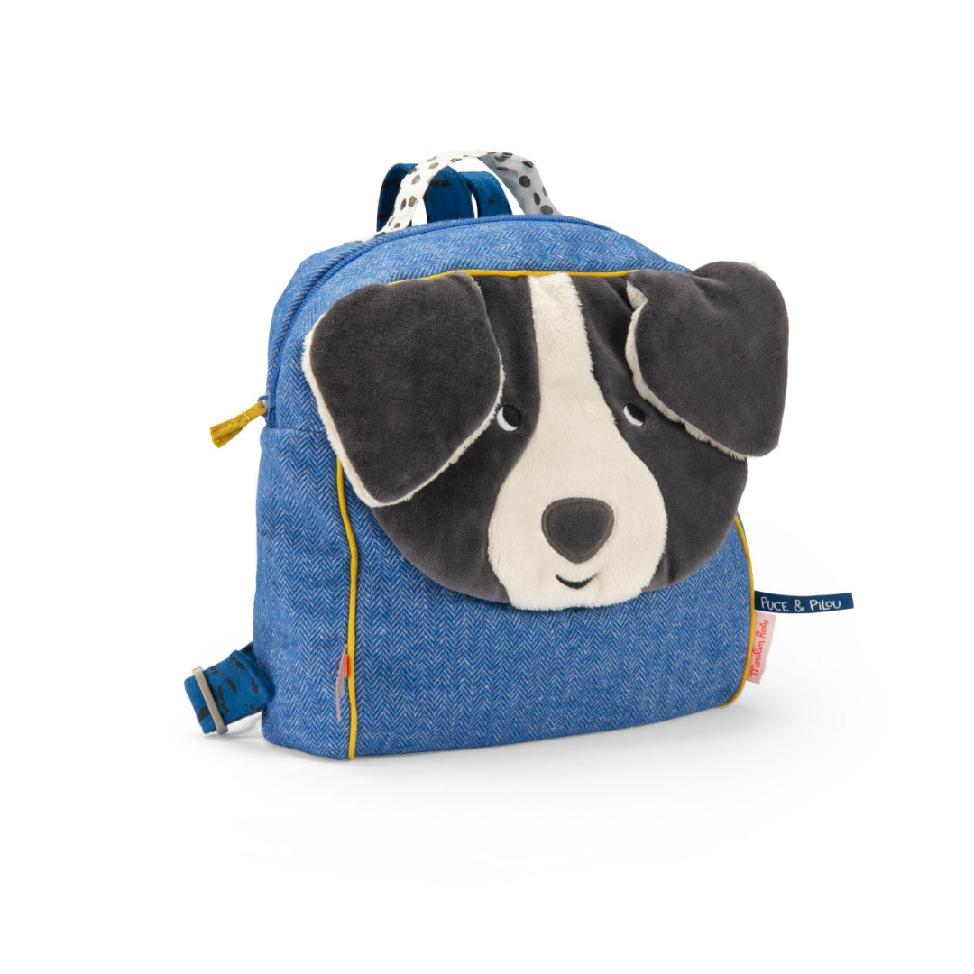 A blue denim-textured dog face backpack by Skip Hop, featuring a plush black and white dog face on the front with floppy ears, yellow piping details, and a small brand tag on the side.