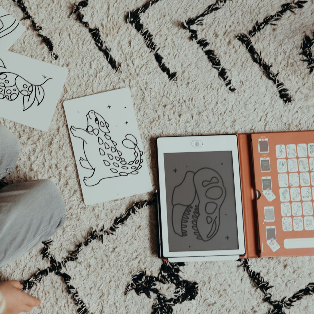 Overhead view of the KIDYDRAW MINI with a T-rex skull drawing, orange card holder and an ankylosaurus card on a cream rug