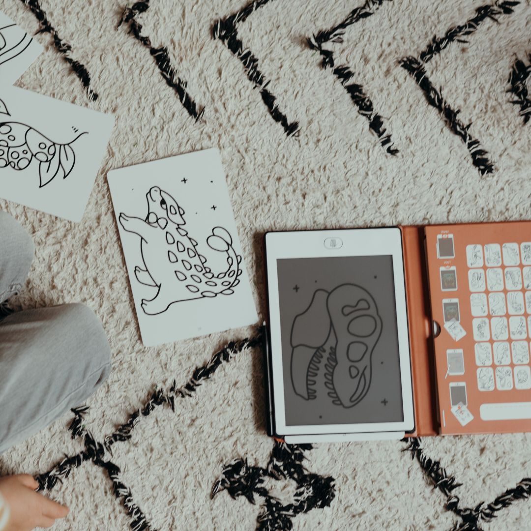 Overhead view of the KIDYDRAW MINI with a T-rex skull drawing, orange card holder and an ankylosaurus card on a cream rug