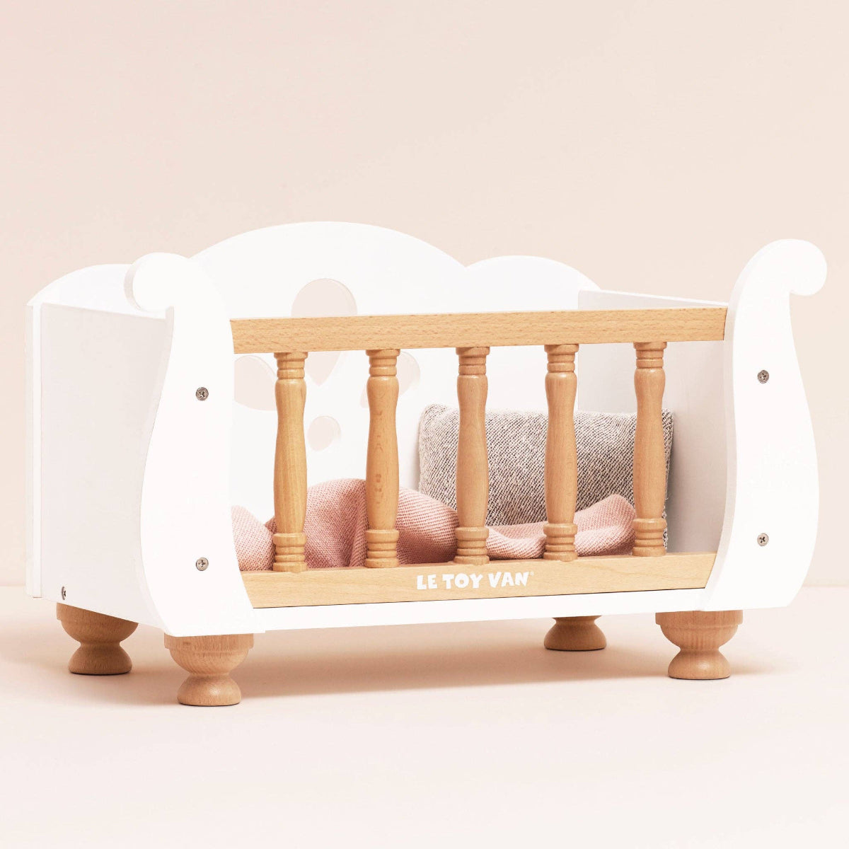 Wooden crib with white mattress and pink pillows on a beige background, featuring Le Toy Van branding.