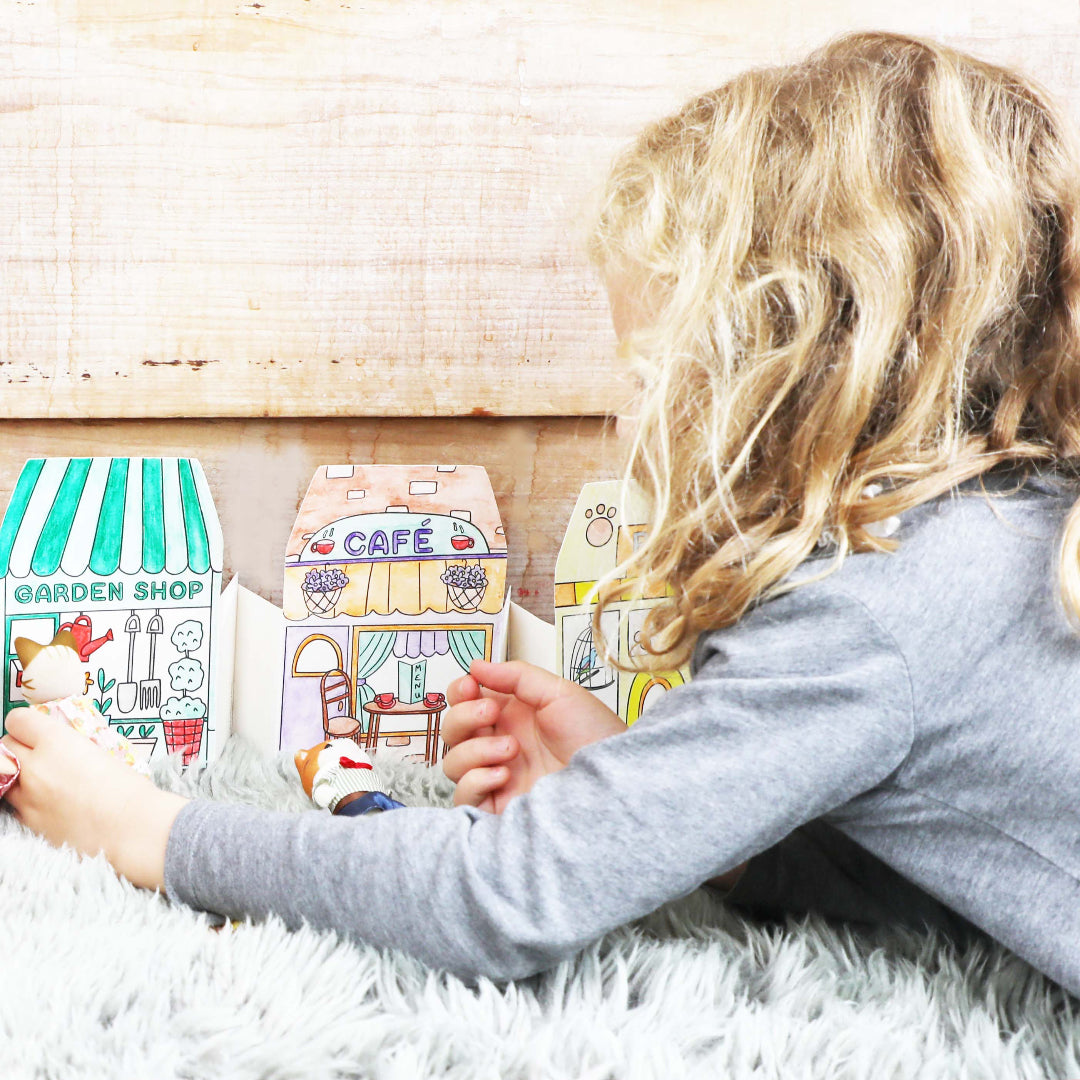 Child playing with toy cardboard houses labeled 'Garden Shop' and 'Café' on a fluffy white rug.