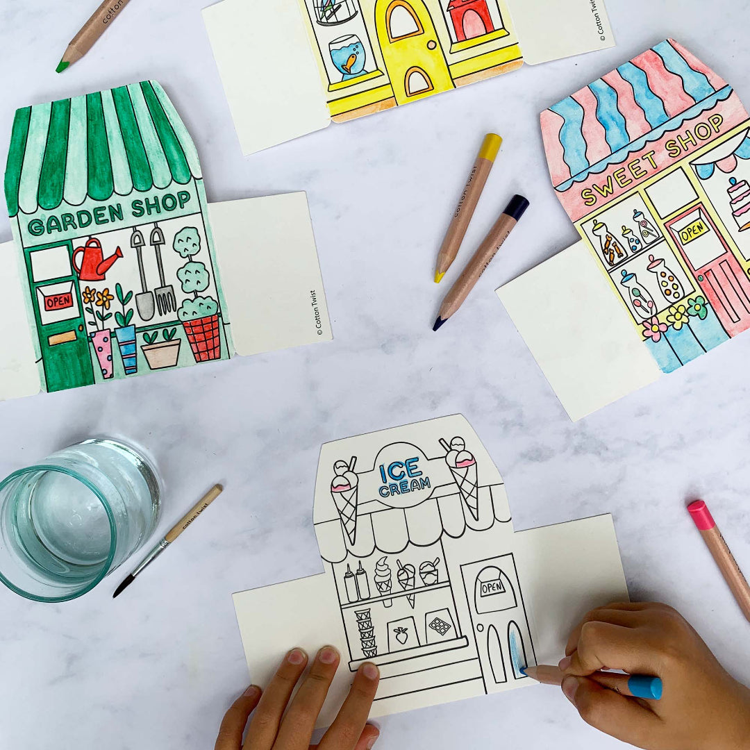Colouring cards with illustrations of shops on a table with colouring supplies.