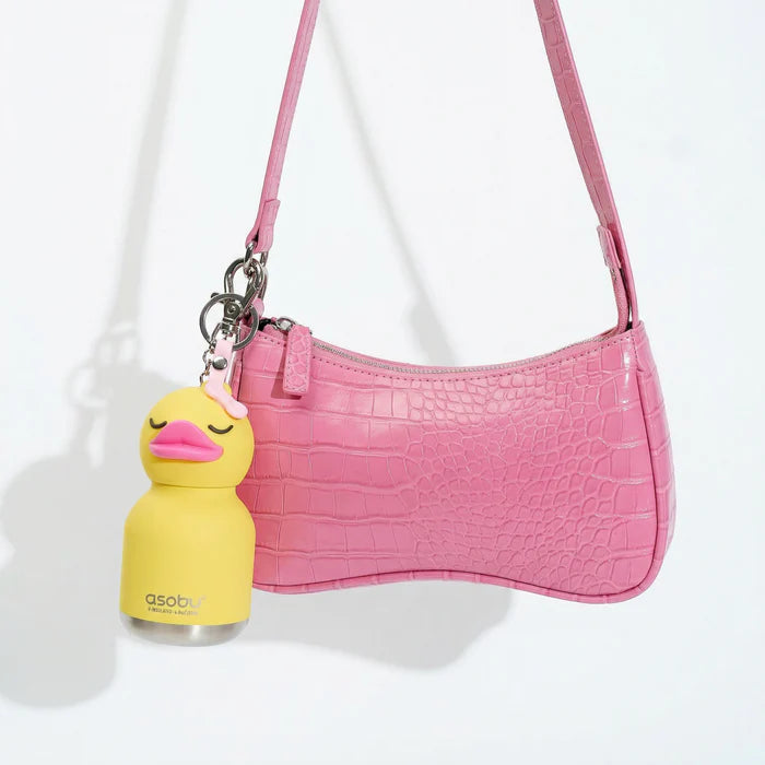 A pink crocodile-embossed shoulder bag with a matching strap, featuring a yellow duck-shaped keychain charm with a small Asobi bottle attached.