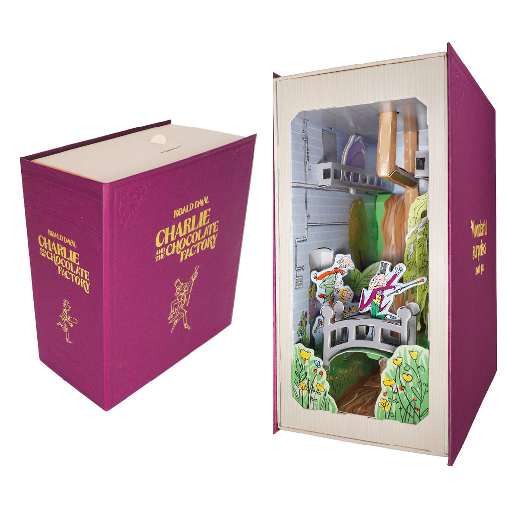 A 3D pop-up cardboard model of 'The Chocolate Factory' set inside a replica magenta book-shaped box. The left side shows the closed book box with gold foil text and illustrations, while the right side shows the open book displaying the detailed pop-up scene with characters and the factory interior.