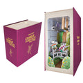 A 3D pop-up cardboard model of 'The Chocolate Factory' set inside a replica magenta book-shaped box. The left side shows the closed book box with gold foil text and illustrations, while the right side shows the open book displaying the detailed pop-up scene with characters and the factory interior.