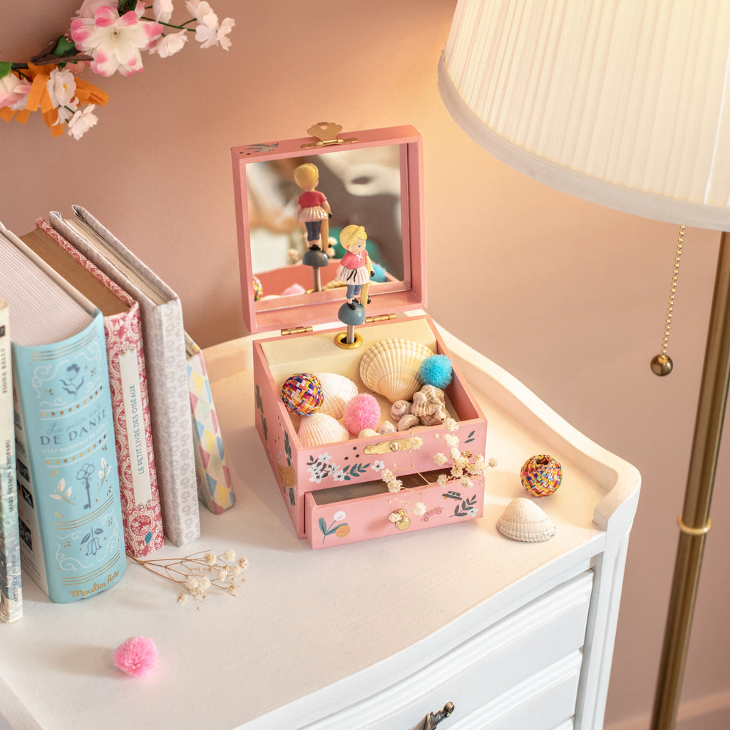Pink jewelry box with decorative items on a dresser next to books and a lamp.