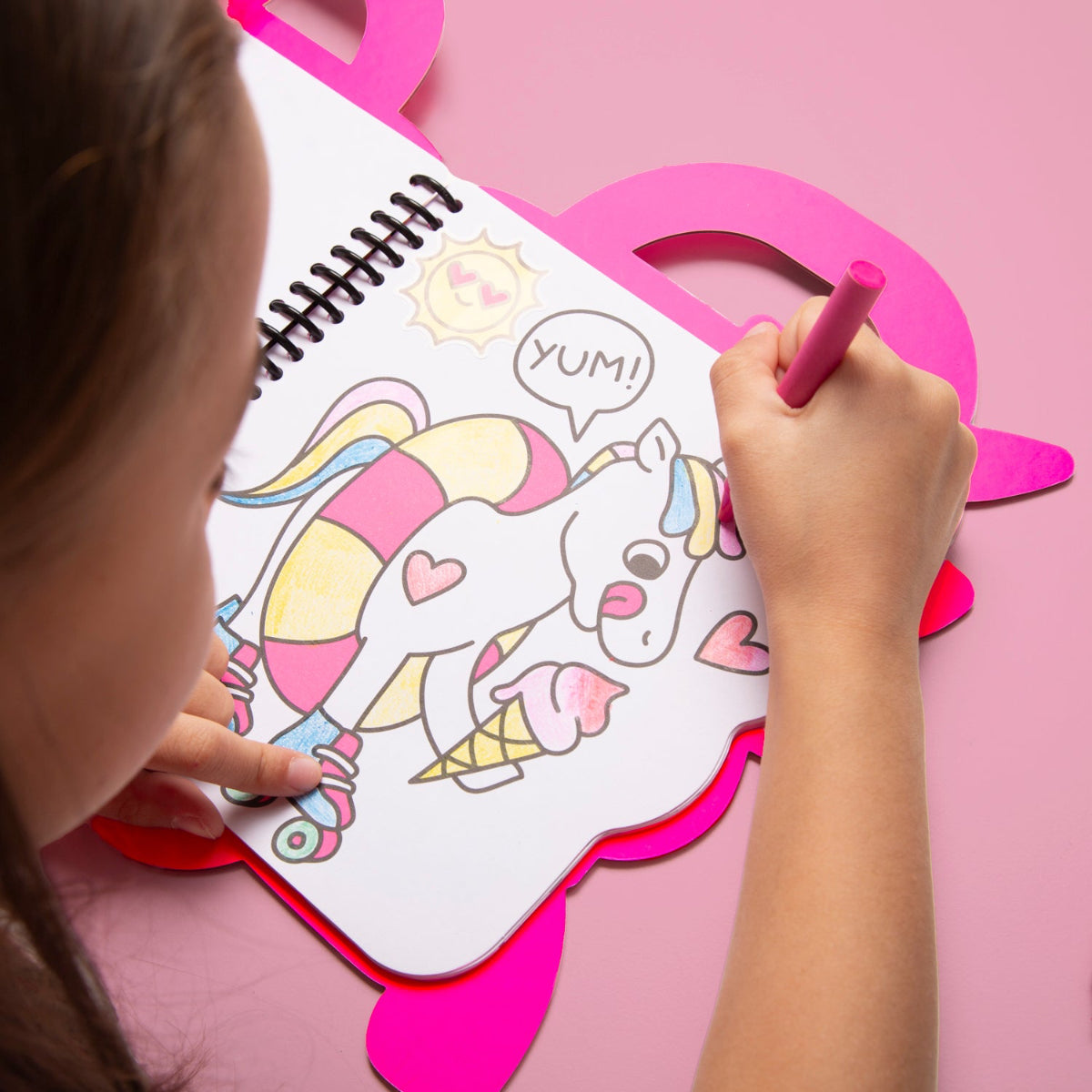 Child coloring a unicorn with pink crayon on a pink heart-shaped stand.
